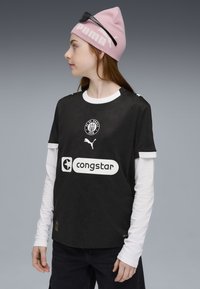 Sort fodboldtrøje med struktureret mønster, hvid krave og trykte logoer. Sammen med en hvid langærmet skjorte under og denimbukser.