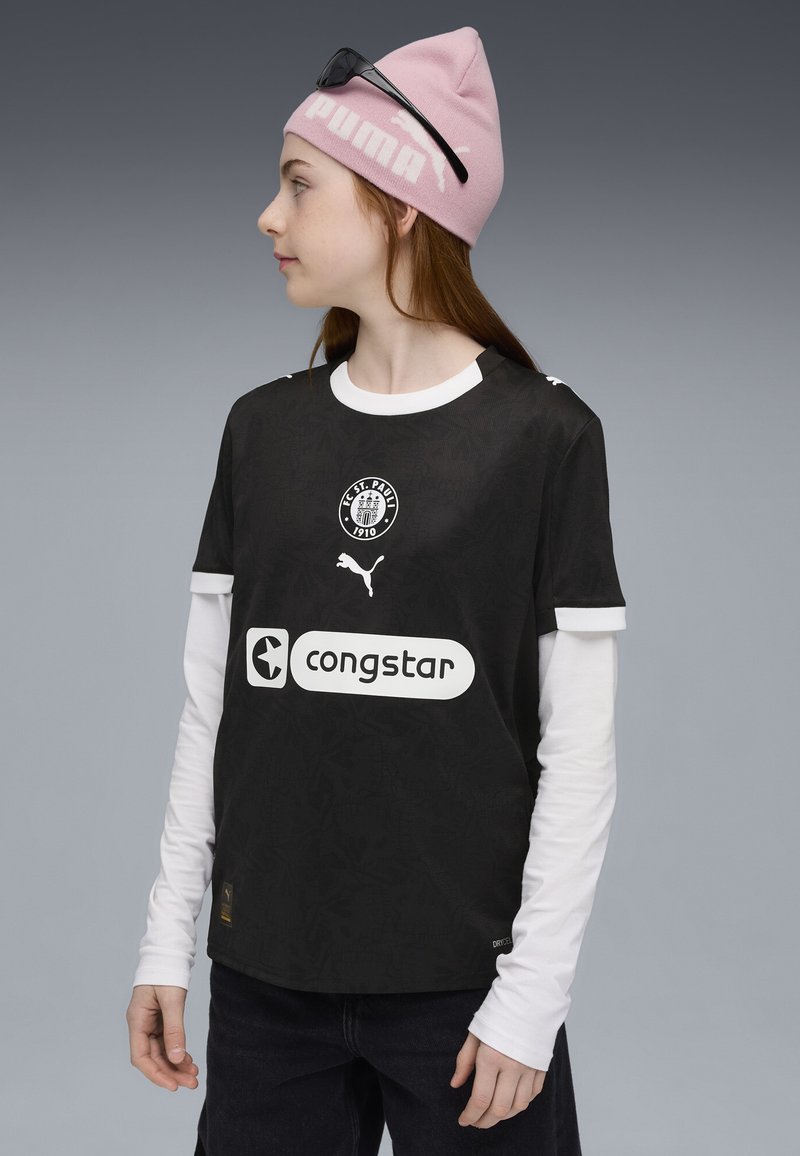 Sort fodboldtrøje med struktureret mønster, hvid krave og trykte logoer. Sammen med en hvid langærmet skjorte under og denimbukser.