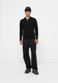 Zwarte longsleeve polo met een ritskraag, gecombineerd met donkere broek en zwarte sportieve schoenen. Het model draagt een donkere pet.