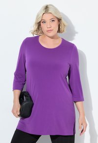 Fialový tunikový top s rukávy po lokty, kulatým výstřihem a bočními rozparky. Nosit s černými kalhotami a držet černou tašku. Hladká textura.
