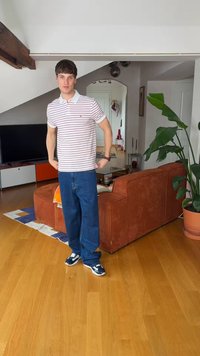 Jeune homme en polo rayé, jean bleu et baskets, debout dans un salon moderne avec un sol en bois, un canapé couleur rouille, une télévision et une grande plante en pot.