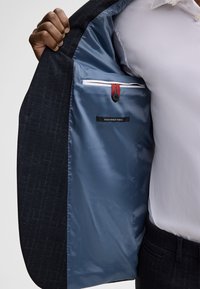 Marineblazer met een gestructureerde buitenstof. Aan de binnenkant heeft het een lichtblauwe voering met een label, een knoop en een zak met rode en witte accenten.