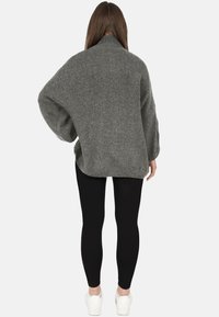 Donna con capelli castani lunghi che indossa un maglione grigio a collo alto e ampio, leggings neri e sneakers bianche, in piedi di schiena.