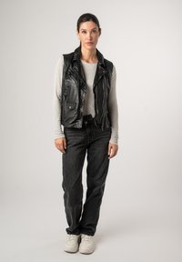 Gilet di pelle nera con revers sopra un maglione grigio chiaro in maglia, abbinato a jeans scuri e sneakers bianche. Il modello è in piedi contro uno sfondo neutro.
