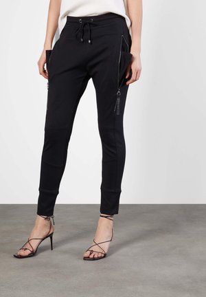 Femme portant un pantalon noir ajusté avec de grandes poches latérales zippées et des sandales noires à talons hauts avec lanières, debout sur un sol gris.
