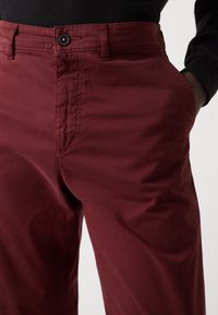 Pantalons bordeaux en tissu lisse avec poches latérales, fermeture à bouton et détails de couture visibles. Coupe ajustée à la taille.