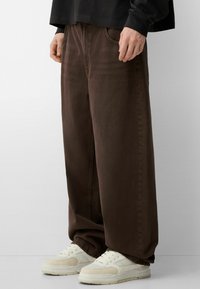 Smešani pamuk s smešnim detaljima, smeđe široke traperice s visokim strukom i opuštenim krojem, u kombinaciji s bež tenisicama.