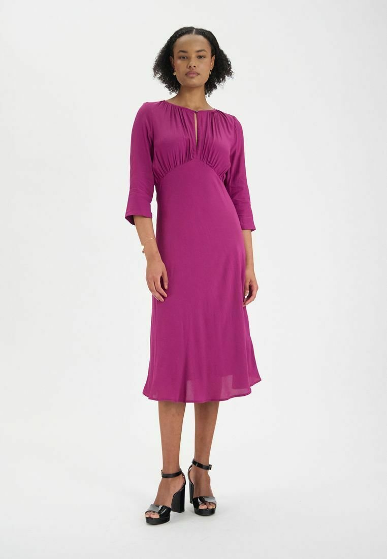 Robe fuchsia, longueur genou, en tissu lisse avec manches trois-quarts et détails de plis pince à l'encolure. Stylée avec des talons noirs épais.