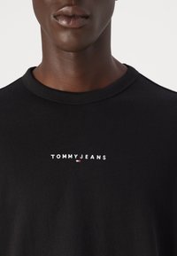 Černé tričko s krátkým rukávem a kulatým výstřihem. Obsahuje bílé vyšívané logo "TOMMYJEANS" a malý červený a modrý akcent pod textem.