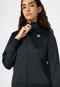 Czarna sportowa kurtka z wysokim kołnierzem, zapinana na zamek z przodu, z elastycznymi mankietami. Posiada małe białe logo na klatce piersiowej oraz kieszenie boczne.