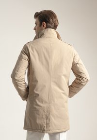 Abrigo beige claro con cuello alto, aberturas traseras y textura suave. Mangas largas y corte recto, diseñado para una silueta ajustada.