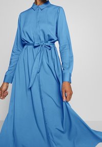 Robe bleu clair à manches longues avec un devant boutonné et une ceinture nouée à la taille. Confectionnée dans un tissu lisse avec une jupe fluide.