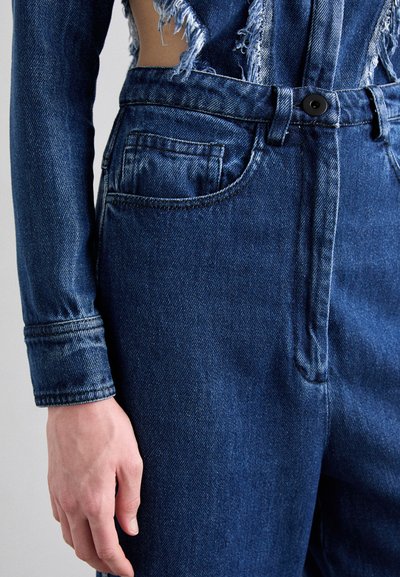 Mørkeblå denim overall med slitte detaljer. Har en frontlomme, beltehemper og en svart knapp i midjen. Glatt tekstur.