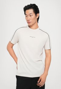 Camiseta de algodón de color beige con mangas cortas, que presenta costuras de acento negras a lo largo de las costuras de los hombros y un pequeño logo de la marca en el centro.