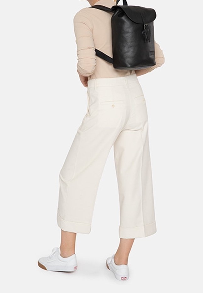Personne portant un haut beige, un pantalon blanc large et court, des baskets blanches et un sac à dos en cuir noir, debout devant un fond clair.