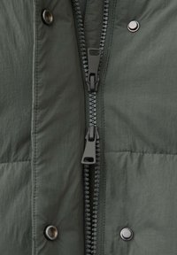 Grüner Pufferjackenstoff mit matter Textur, ausgestattet mit einem doppelten Reißverschluss und Druckknopfverschlüssen. Auffällige schwarze Hardwareakzente zusammen mit Nahtdetails.