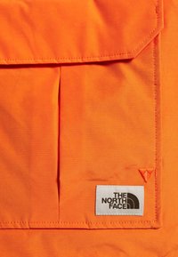 Světlý oranžový textil s klopovou kapsou a štítkem s brandingem "The North Face". Hladká textura s kontrastním šitím a detaily loga.