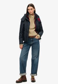 Doudoune marine avec des accents rouges, pull beige texturé, manches retroussées sur un jean bleu, et bottes marron à lacets.