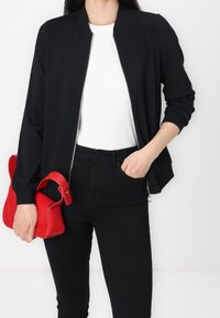 Persona che indossa una giacca nera con zip, una maglietta bianca a costine, pantaloni neri, tiene una borsa rossa con una mano e l'altra mano in tasca.