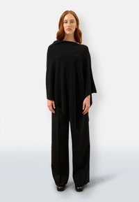 Czarny poncho wykonane z miękkiego materiału, charakteryzujące się zwiewnym krojem w kształcie trójkąta, noszone na czarnych szerokich spodniach.