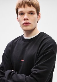 Μαύρη μπλούζα crewneck με ριμπ μανσέτες και στρίφωμα. Διαθέτει μικρό κόκκινο λογότυπο "HUGO" στο στήθος. Το μοντέλο φοράει από κάτω ένα άσπρο T-shirt.