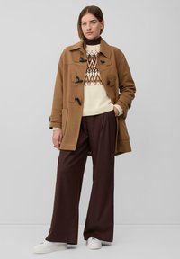 Vrouw in een camel toggle coat over een gemusterd crèmekleurig truitje, brede bruine broek en witte sneakers, staand tegen een effen grijze achtergrond.