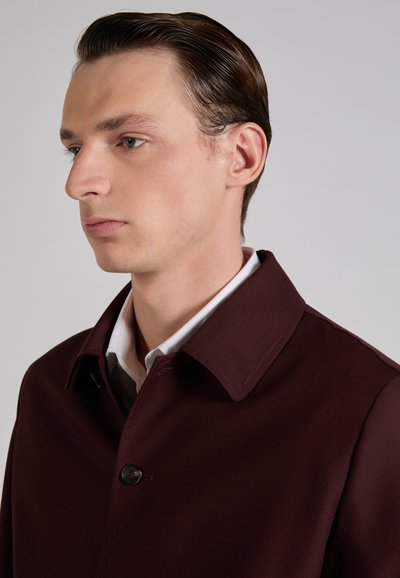 Jeune homme aux cheveux coiffés en arrière portant un manteau bordeaux foncé par-dessus une chemise blanche à col, regardant vers la gauche sur un fond gris uni.