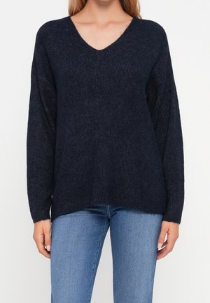 Femme portant un pull en tricot bleu marine à encolure en V et un jean bleu clair, debout devant un fond blanc uni.