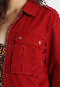 Rode ritsjack met een puntige kraag, twee voorzakken en messing drukknoopjes. Het materiaal is soepel met een lichte textuur.