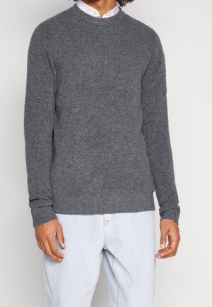 Homme portant un pull en laine gris sur une chemise blanche et un jean bleu clair, debout devant un fond clair uni.