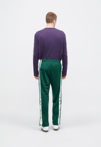 Μωβ πουκάμισο με μακριά μανίκια σε συνδυασμό με πράσινο παντελόνι φόρμας με λευκές ρίγες στα πλάγια και κουμπιά κατά μήκος των ποδιών, με λευκά αθλητικά παπούτσια.