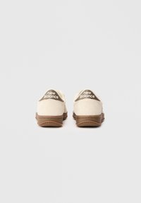 Sneakers New Balance cechują się miękkim beżowym zamszowym wierzchem, brązową gumową podeszwą i białym akcentem logo na pięcie. Gładka tekstura z minimalnym zdobieniem.