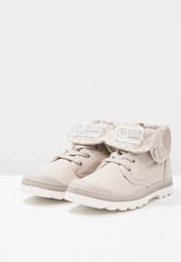 Stivali alla caviglia in tela beige con suole in gomma, caratterizzati da un polsino decorato e dettagli con logo. Presentano lacci e una suola bianca in contrasto.