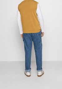 Gul ærmeløs sweater over en hvid langærmet skjorte kombineret med blå cargobukser. Hvide sko fuldender outfittet.