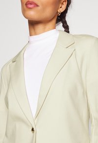 Femme portant un blazer crème sur un haut blanc à col montant, cheveux tressés et petites boucles d'oreilles en forme d’anneau en or.