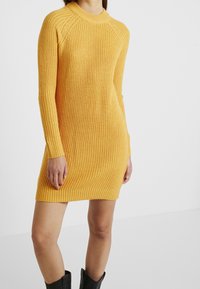 Gelbes gestricktes Kleid mit gerippter Textur, langen Ärmeln und einem runden Ausschnitt. Es passt eng und fällt oberhalb des Oberschenkels.