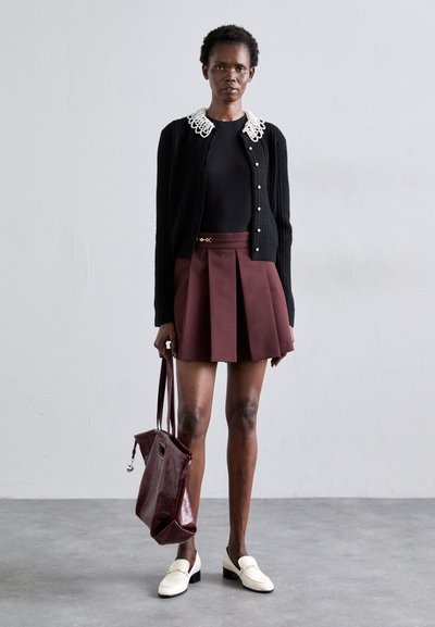 Gilet noir côtelé avec col en dentelle, top noir en dessous, jupe plissée bordeaux, mocassins blancs et sac à main bordeaux brillant.