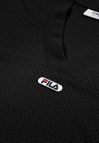 Lähikuva mustasta perforoidusta urheilupaidasta, jossa on V-kaula-aukko ja FILA-logopläkki kauluksen alapuolella keskellä.