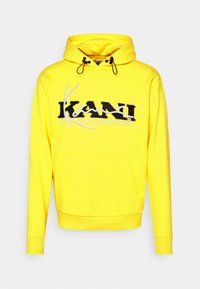 Sudadera amarilla con un logo "KANI" en blanco y negro en el pecho. Cuenta con un bolsillo frontal, capucha con cordón y puños acanalados.