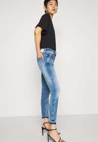 Frau steht in blauen Skinny Jeans, schwarzem kurzärmligen Oberteil und schwarzen High-Heels vor einem einfarbigen hellen Hintergrund.