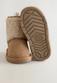 Braune Wildlederstiefelette mit einem flauschigen beigen Obermaterial, strukturierten Gummisohle mit einem Zickzackmuster und dekorativem Strickzugband.