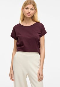 Burgundska bombažna majica z okroglim izrezom in zavitimi rokavi, v kombinaciji s kremnimi širokimi hlačami, prikazana na nevtralni podlagi.