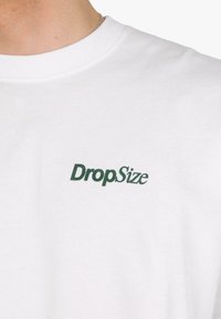 T-shirt en coton blanc avec un logo "DropSize" vert sur la poitrine gauche. Col rond avec texture côtelée.