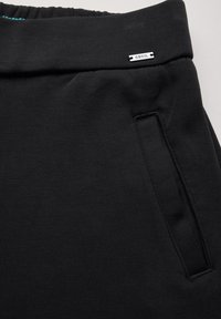 Un pantalon de survêtement noir avec une taille élastique, une poche latérale et une petite étiquette de marque. Le tissu a une texture lisse et est légèrement extensible.