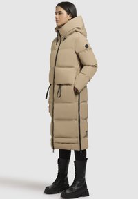 Beige pufferjakke med høj krave, to sidelommer, sorte lynlåse og design i knælængde. Parret med sorte snørebåndsstøvler.