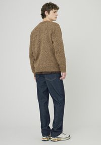 Pull en maille marron à motifs tachetés avec des poignets côtelés, associé à un jean en denim foncé. Les baskets ont une semelle blanche et dorée, accentuant l'ensemble.