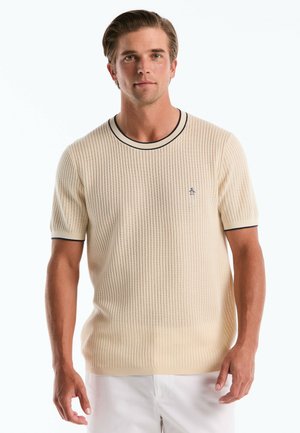 Man met lichtbruin haar die een beige geribbelde korte mouwtrui met zwarte rand en witte broek draagt, tegen een witte achtergrond.