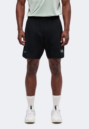 Homme portant un short noir EA7, des chaussettes blanches, des baskets blanches et un t-shirt gris clair, debout avec les bras détendus le long du corps.