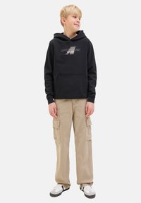 Sweatshirt à capuche noire avec poche avant et logo. Pantalons cargo beiges avec poches latérales. Baskets blanches et noires. Fond uni intérieur.