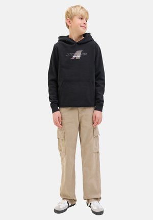 Zwarte hoodie met voorzak en logo. Beige cargo broek met zijzakken. Wit en zwarte sneakers. Effen achtergrond binnenshuis.